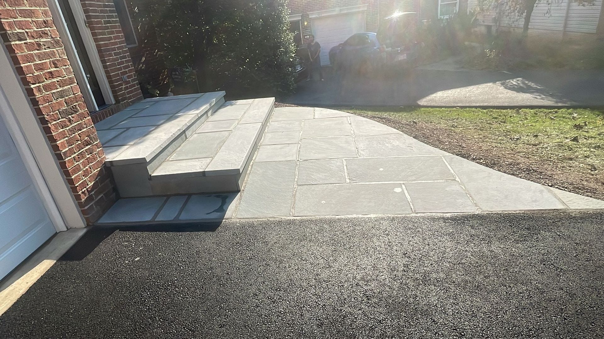 Stone steps and walkway leading to a house entrance, next to asphalt driveway and green grass.