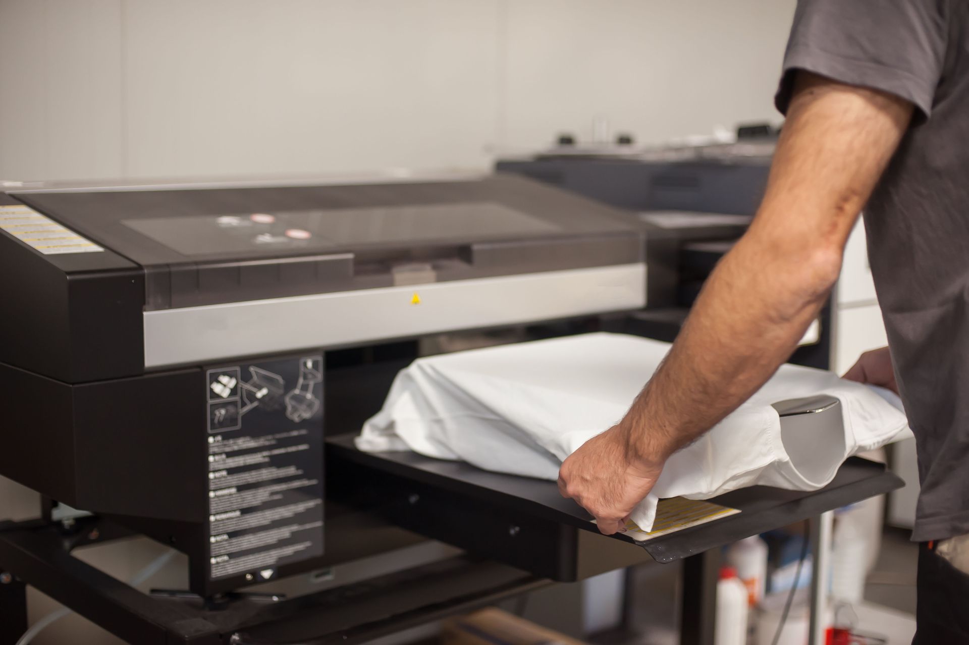 Person placing a white t-shirt on a direct-to-garment printer. The machine is black, in a workshop setting.