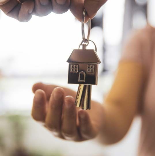 Hand passing house-shaped key to another hand, suggesting a real estate transaction.