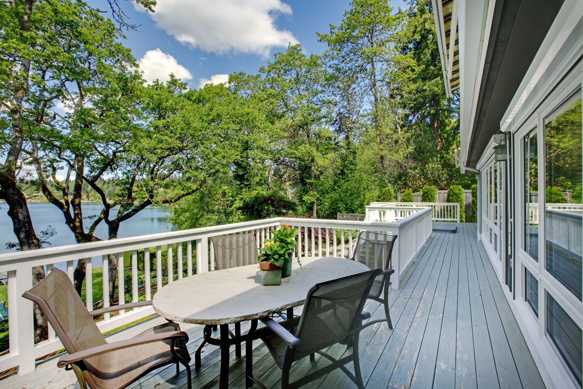 A gray wooden deck with outdoor patio furniture overlooking a lake surrounded by lush green trees under a blue sky.