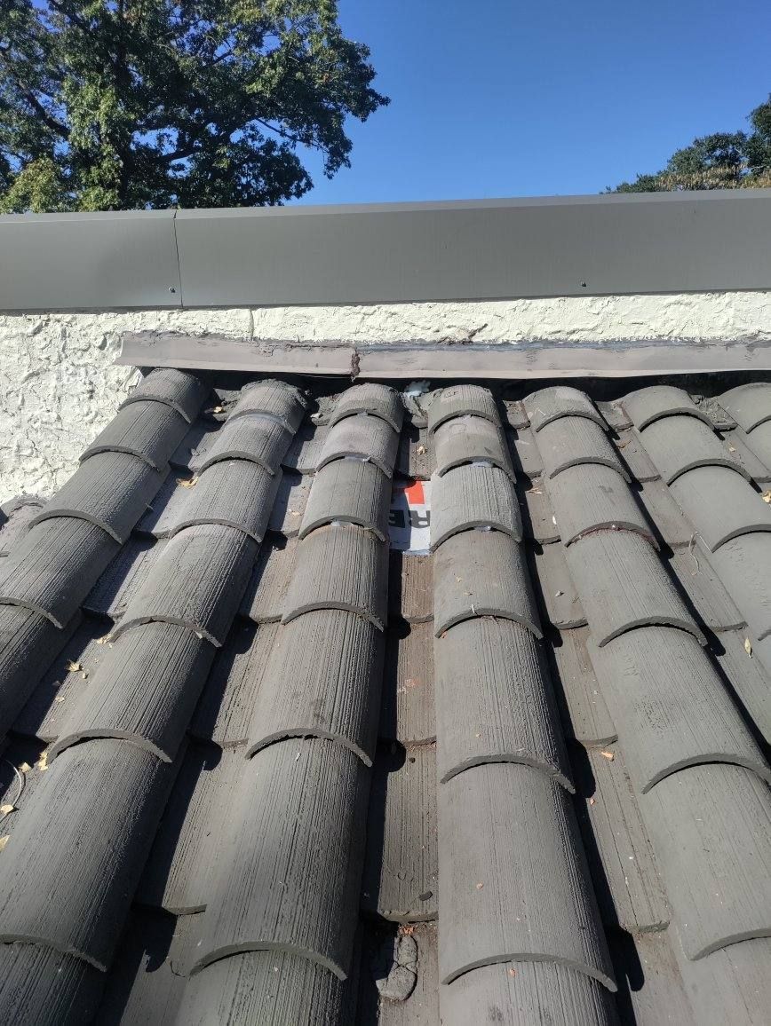 Close-up of a tiled roof, looking upward towards a dark eave. Sky and tree visible in the background.