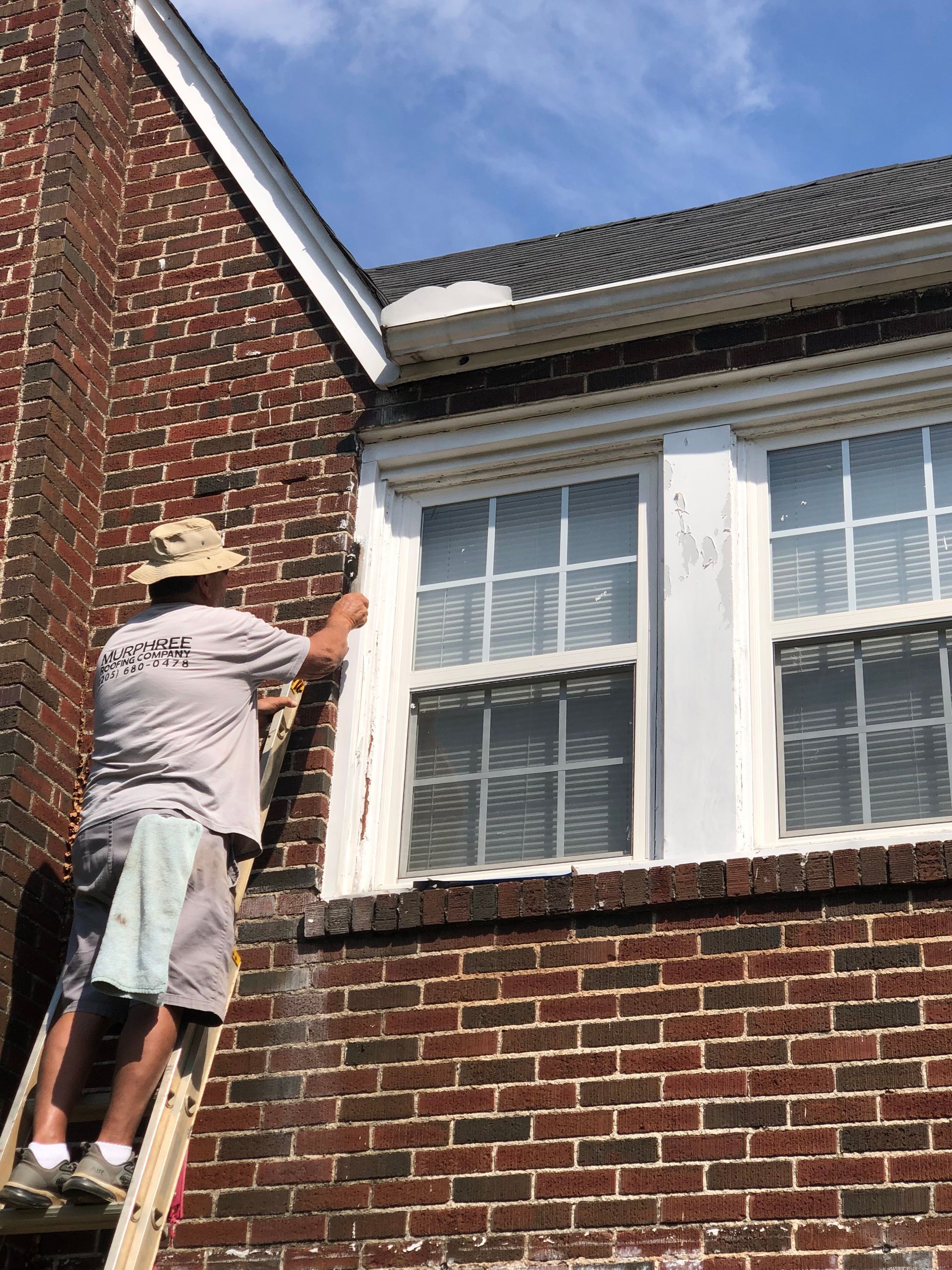 Person on a ladder painting white window trim on a brick building under a blue sky.