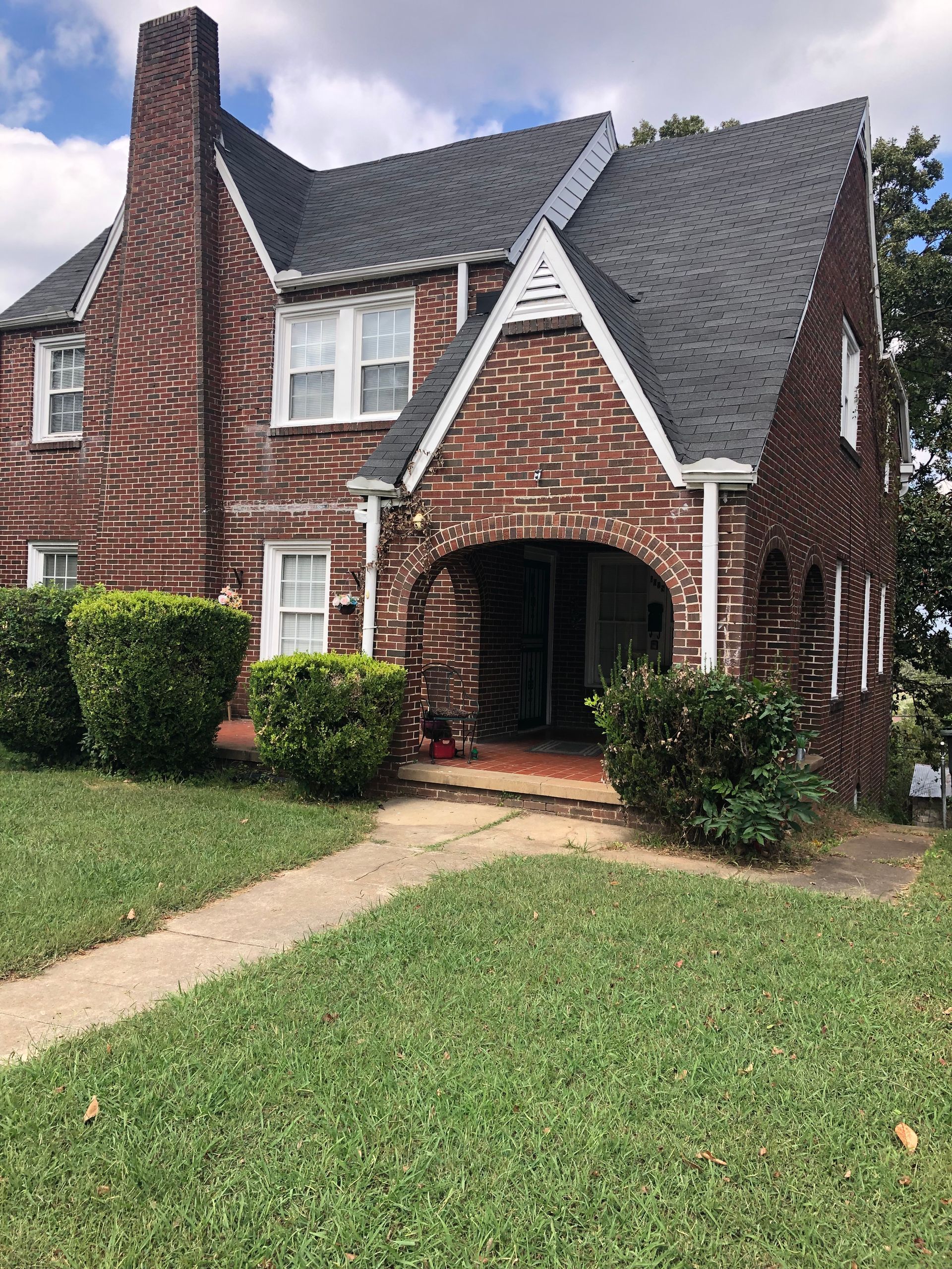 Brick house with arched porch, gabled roof, and chimney. Green lawn and bushes in front.