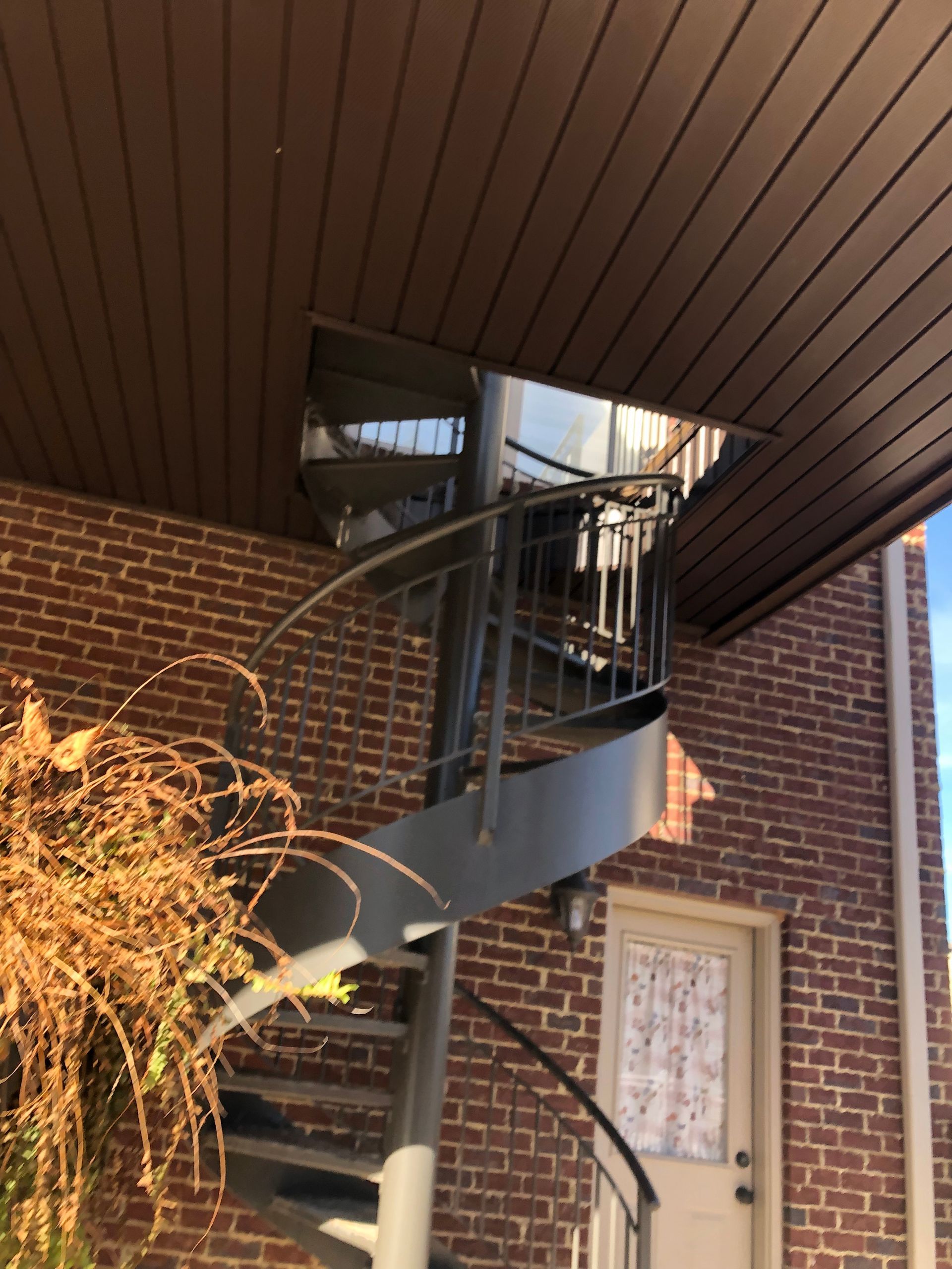 Spiral metal staircase on brick building leading to an opening in the brown overhead.
