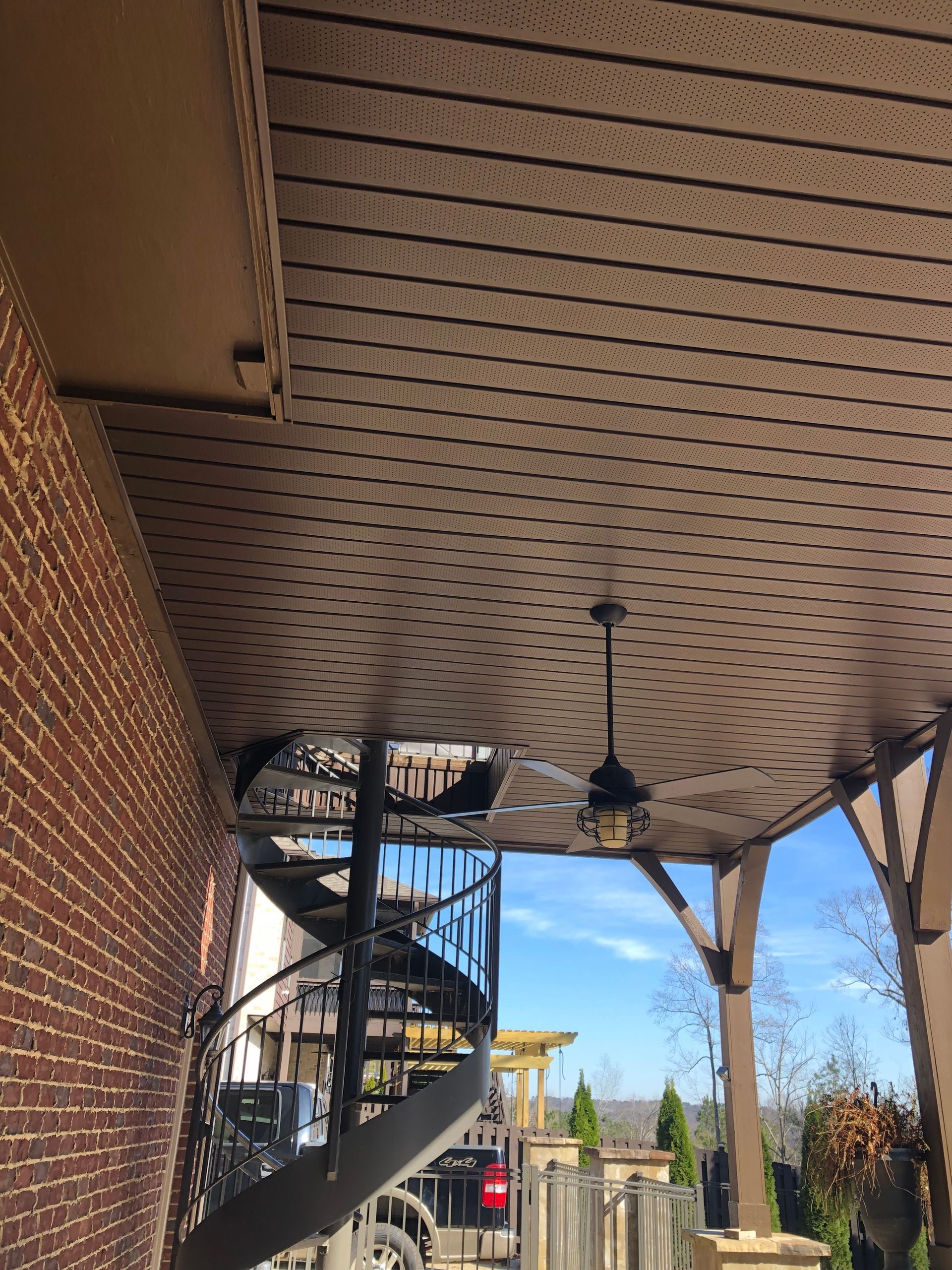 View of a brown, perforated ceiling with a hanging lamp, a spiral staircase, and brick wall. Sunny day.