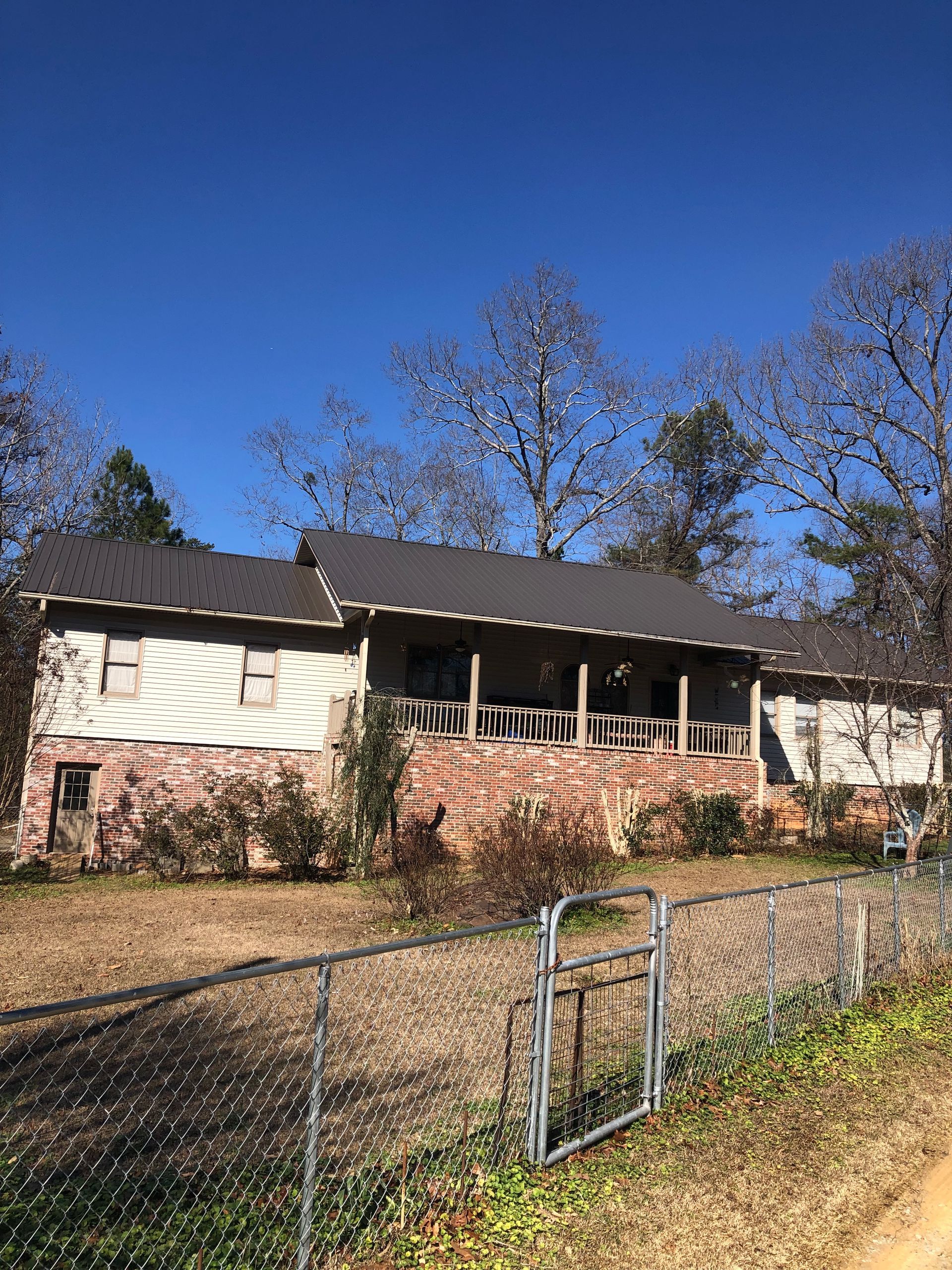 House with a brick base and brown metal roof, porch, and chain-link fence on a sunny day.