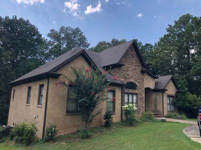 Tan stucco house with stone accents, brown roof, and lush green landscaping.