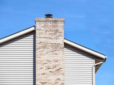 Brick chimney atop a house with beige siding against a clear blue sky.