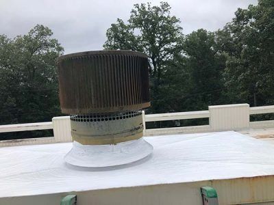 A rooftop vent with a weathered metal top, on a white roof, trees in the background.