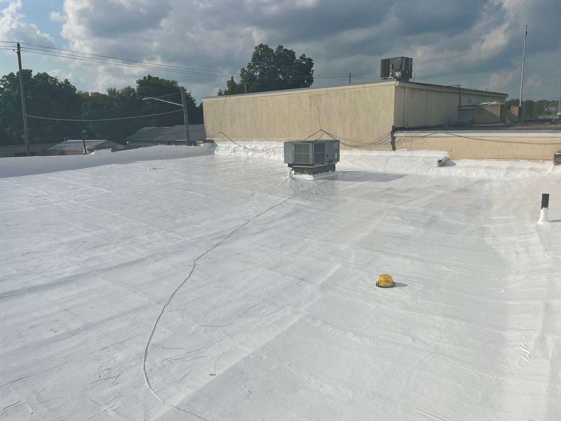 White foam roofing on a commercial building rooftop with a central HVAC unit and surrounding trees under a cloudy sky.