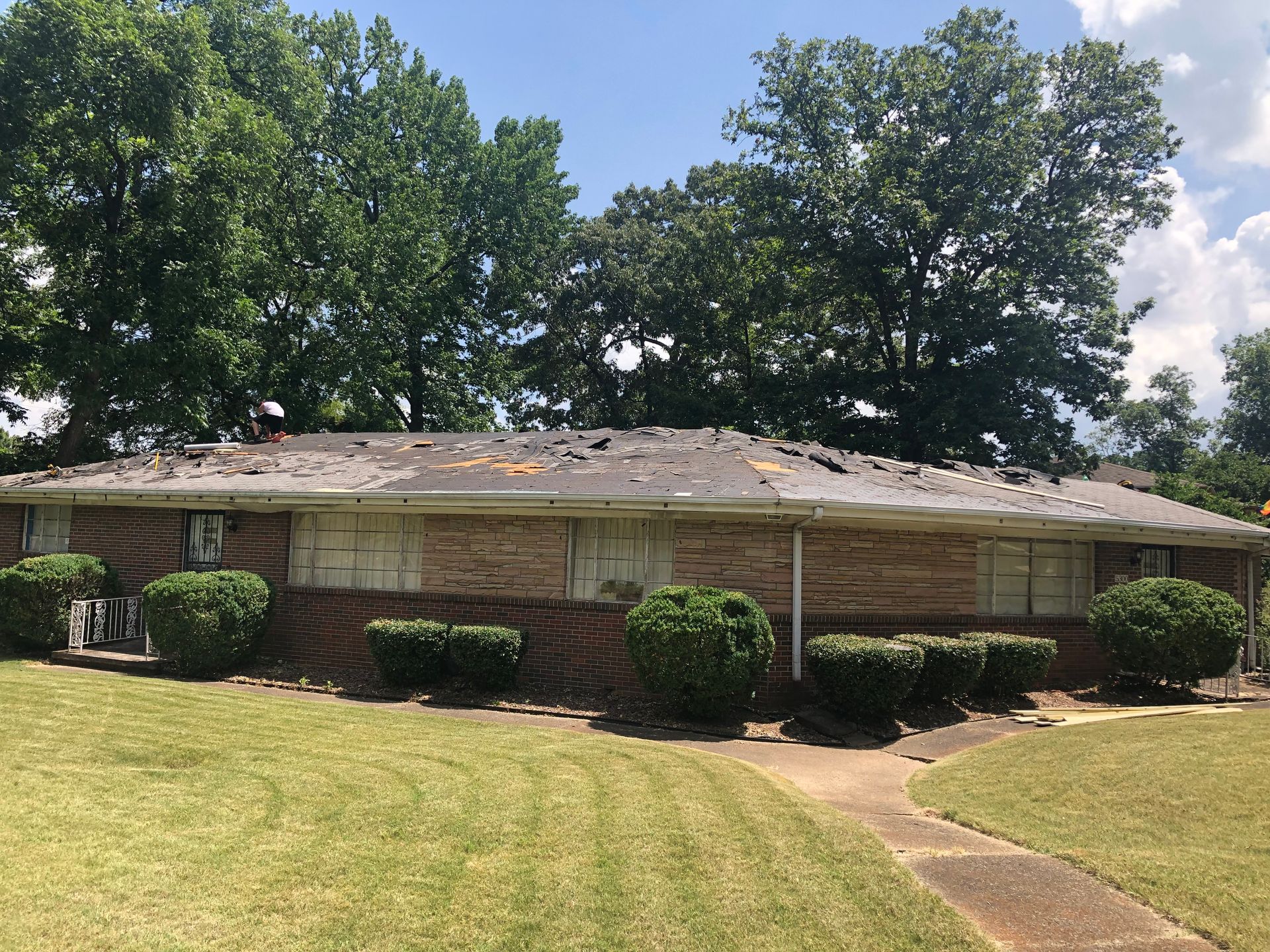 A brick building with a damaged roof and trimmed bushes in front. Green grass and trees are in the background.