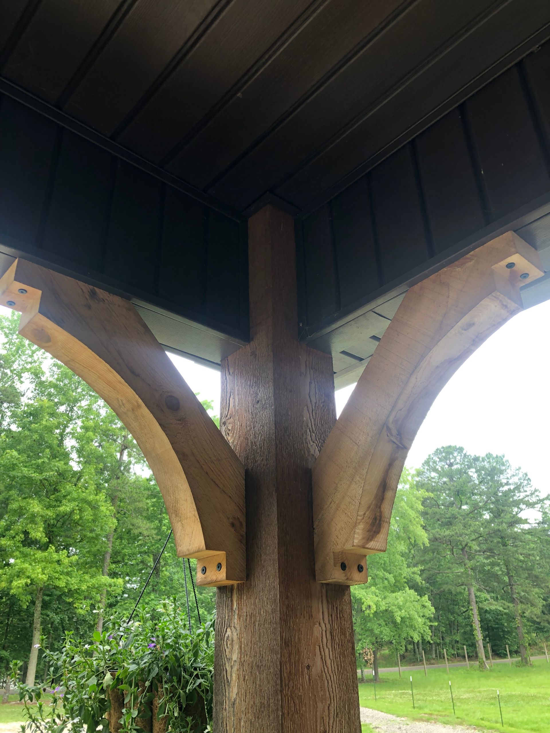 Wooden post with curved brackets supporting a dark ceiling. Outdoors with green trees and grass in the background.
