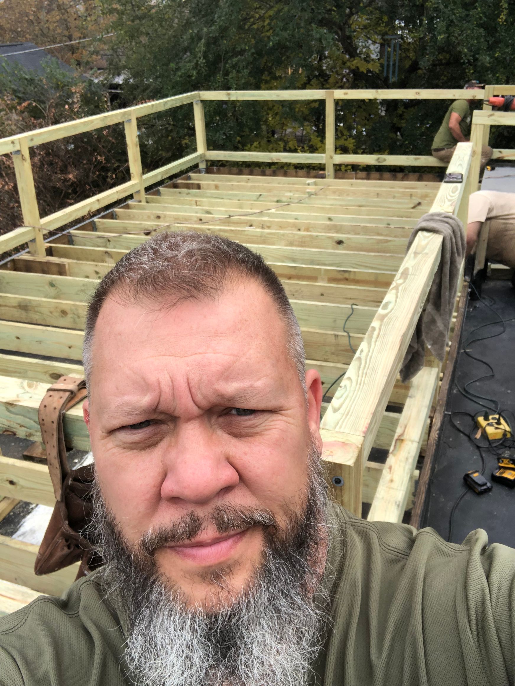 Man with beard taking selfie on unfinished wooden deck with a worker and tools visible.