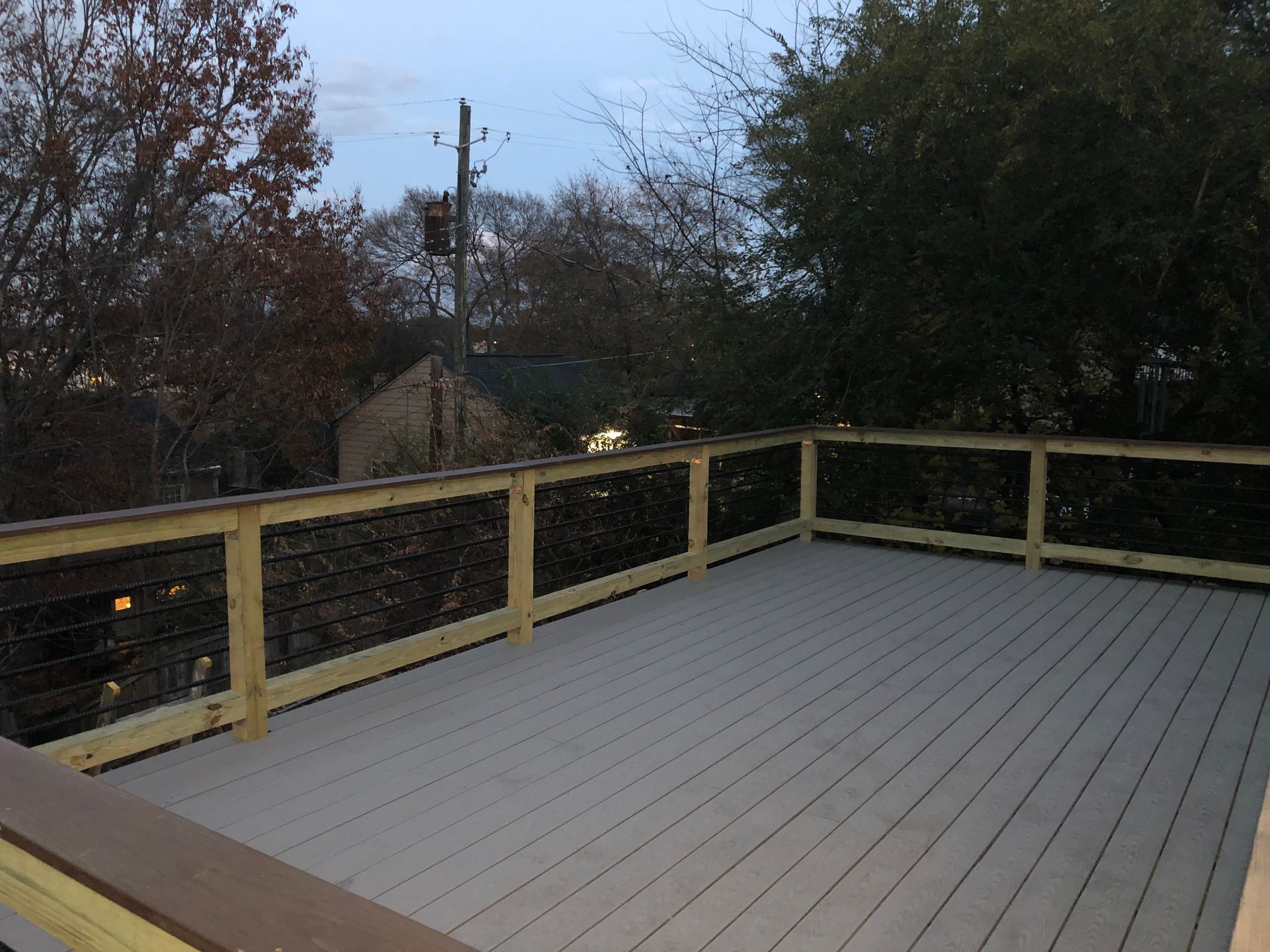 Deck with cable railing, light-colored deck boards, surrounded by trees and a building in the background.