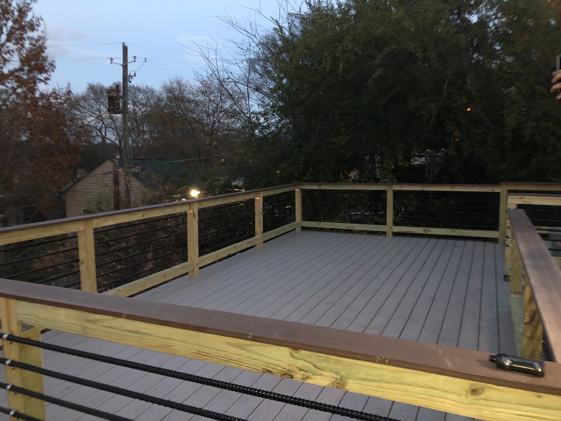 Wooden deck with gray flooring, surrounded by a wire and wood railing. Trees and a building are in the background.