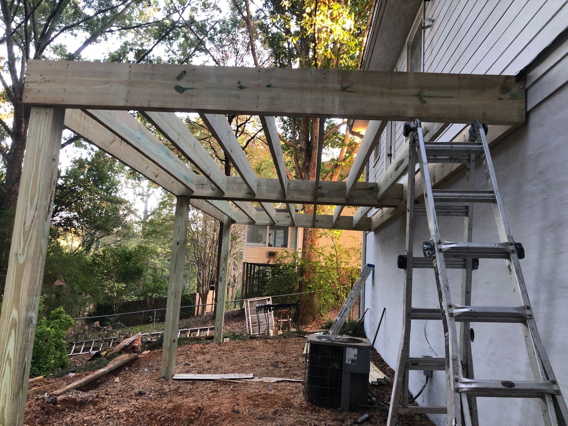 Wooden pergola under construction next to a house with a ladder propped against it.