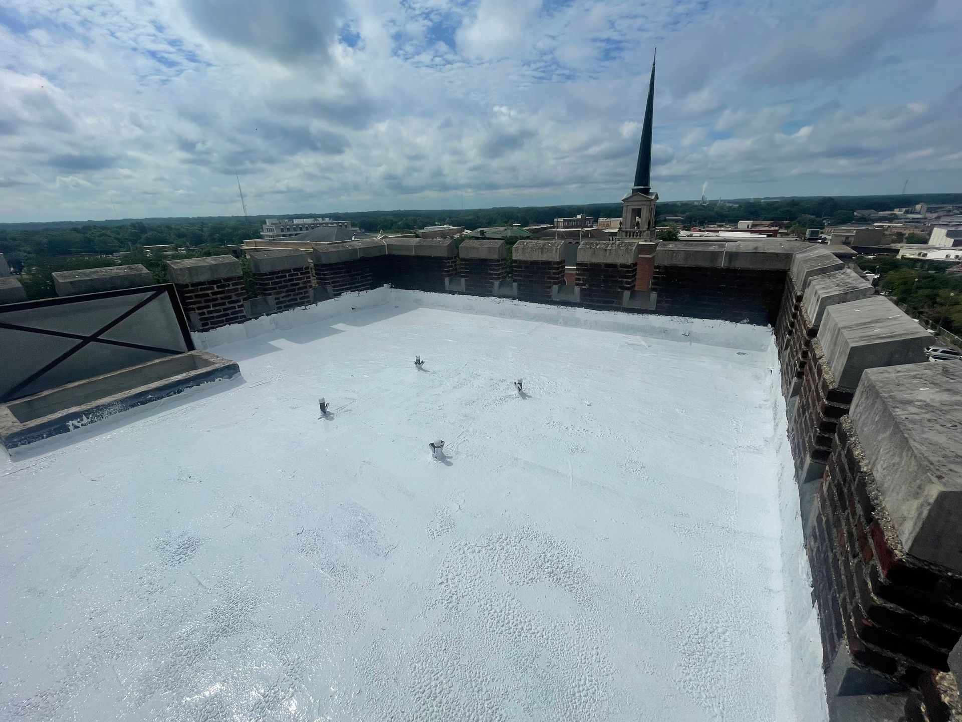View of a flat white roof with metal spikes, part of a brick building. In the distance, a spire and cityscape.