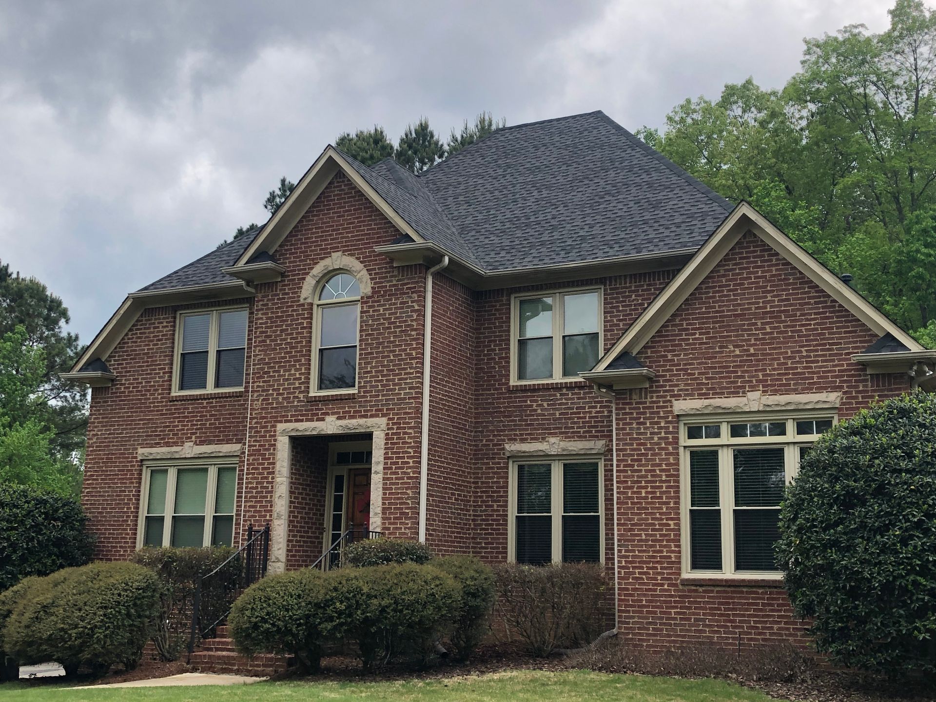 Brick two-story house with dark roof and trim, several windows and bushes in front.