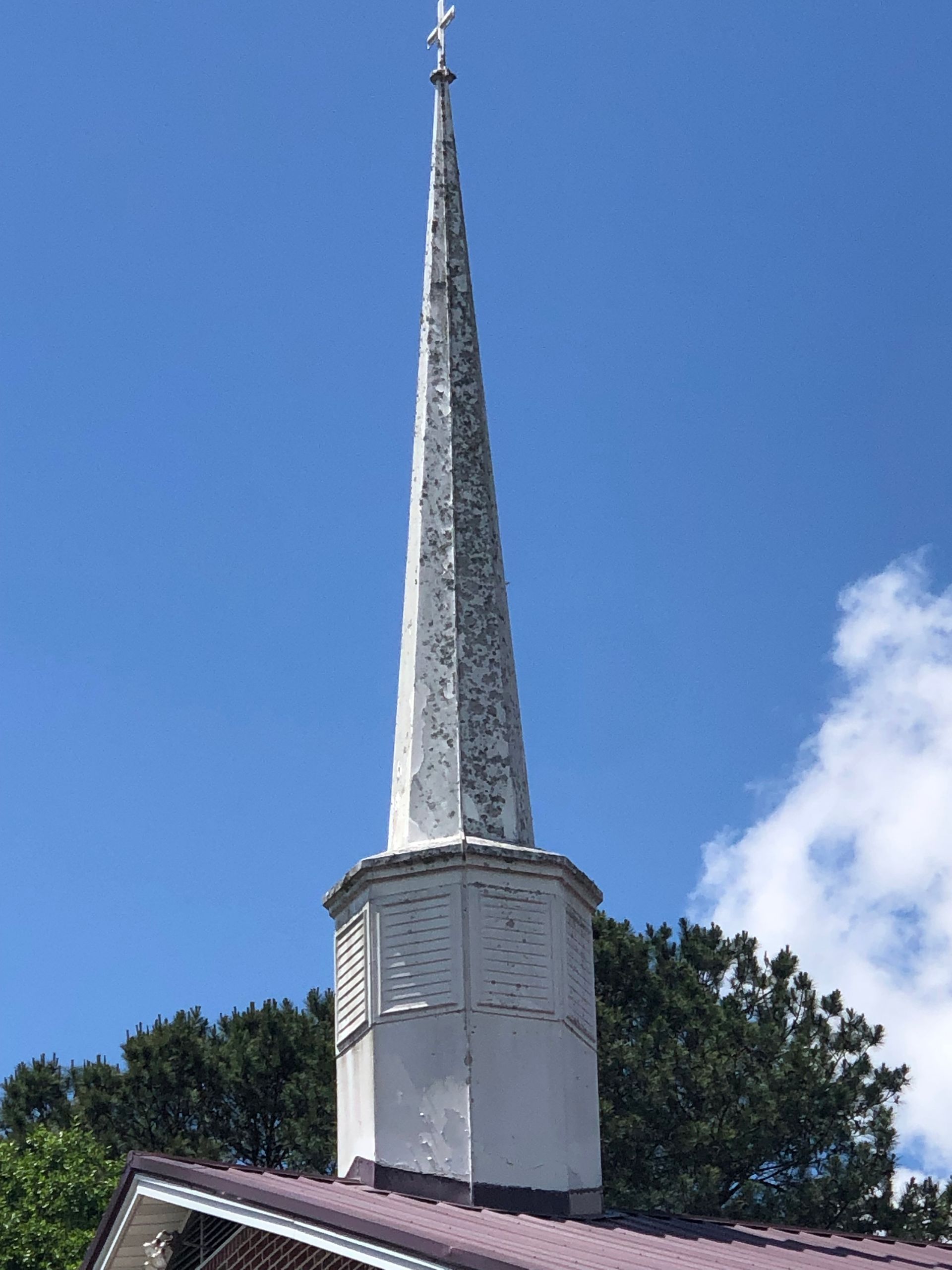White church steeple against a blue sky, topped with a cross. Green trees at base.