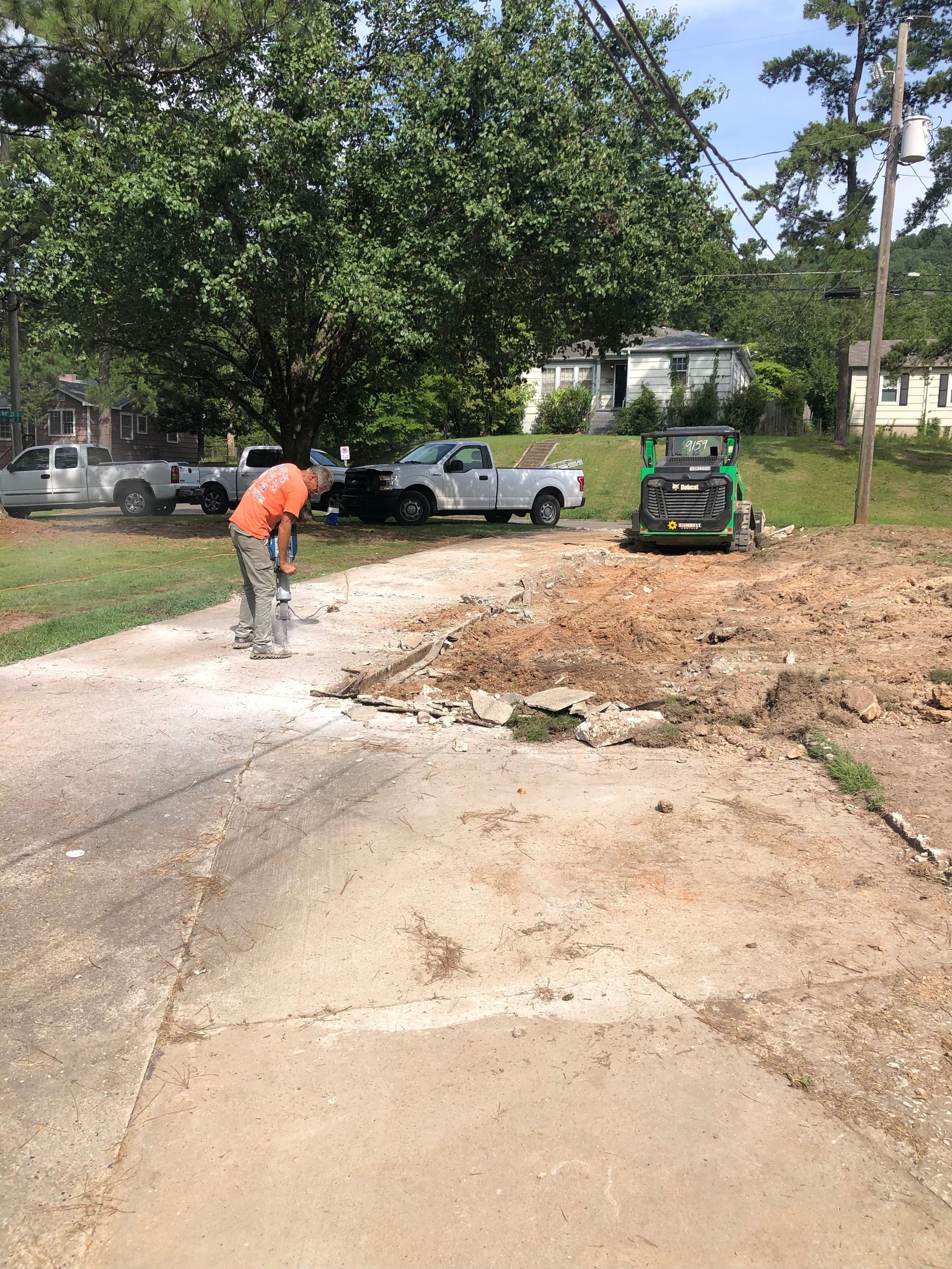Construction worker jackhammering concrete driveway next to parked trucks and a Bobcat, in a residential area.