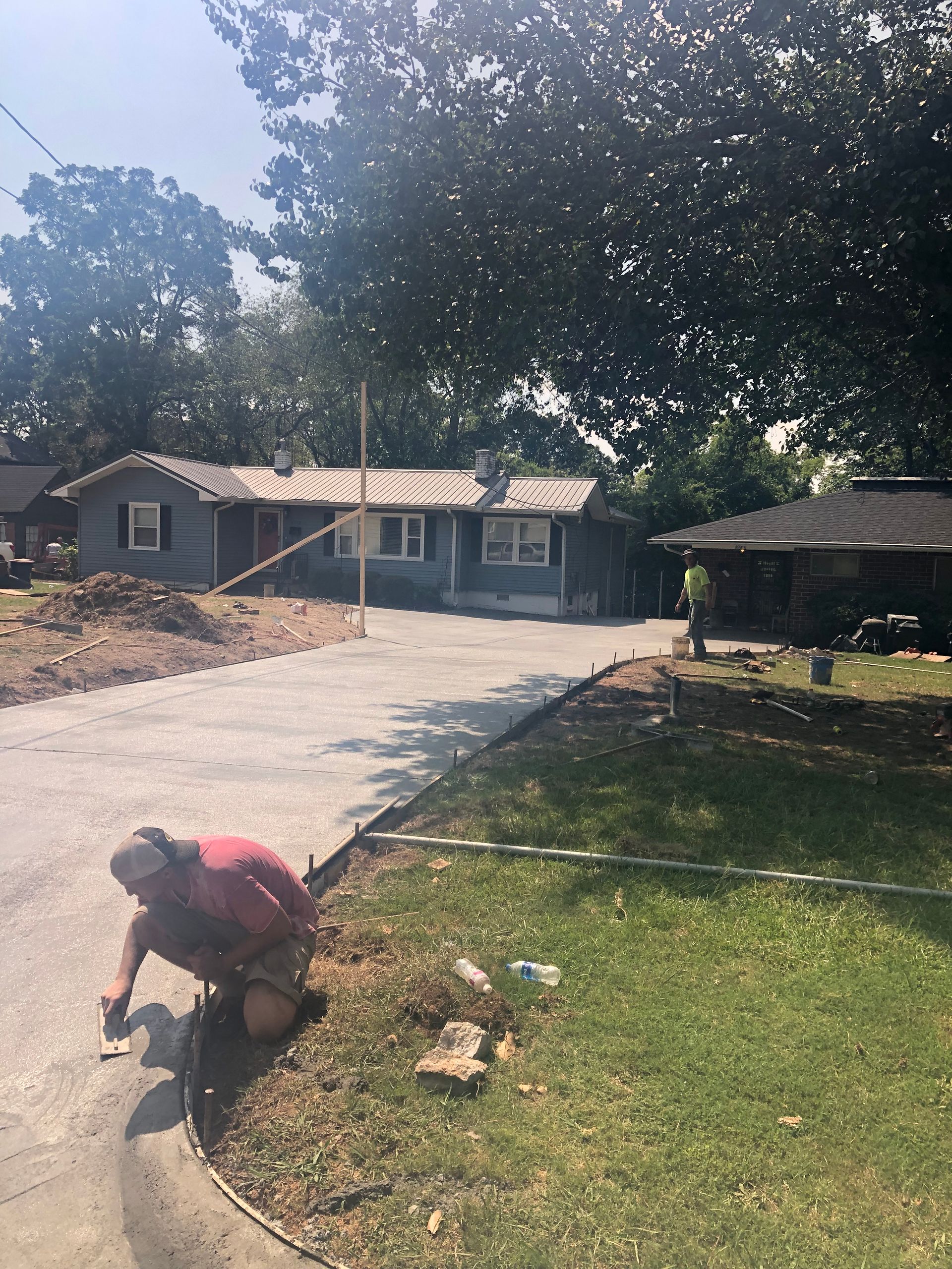 Person kneeling on the ground by curb, working on landscaping with house in the background. Sunny day.