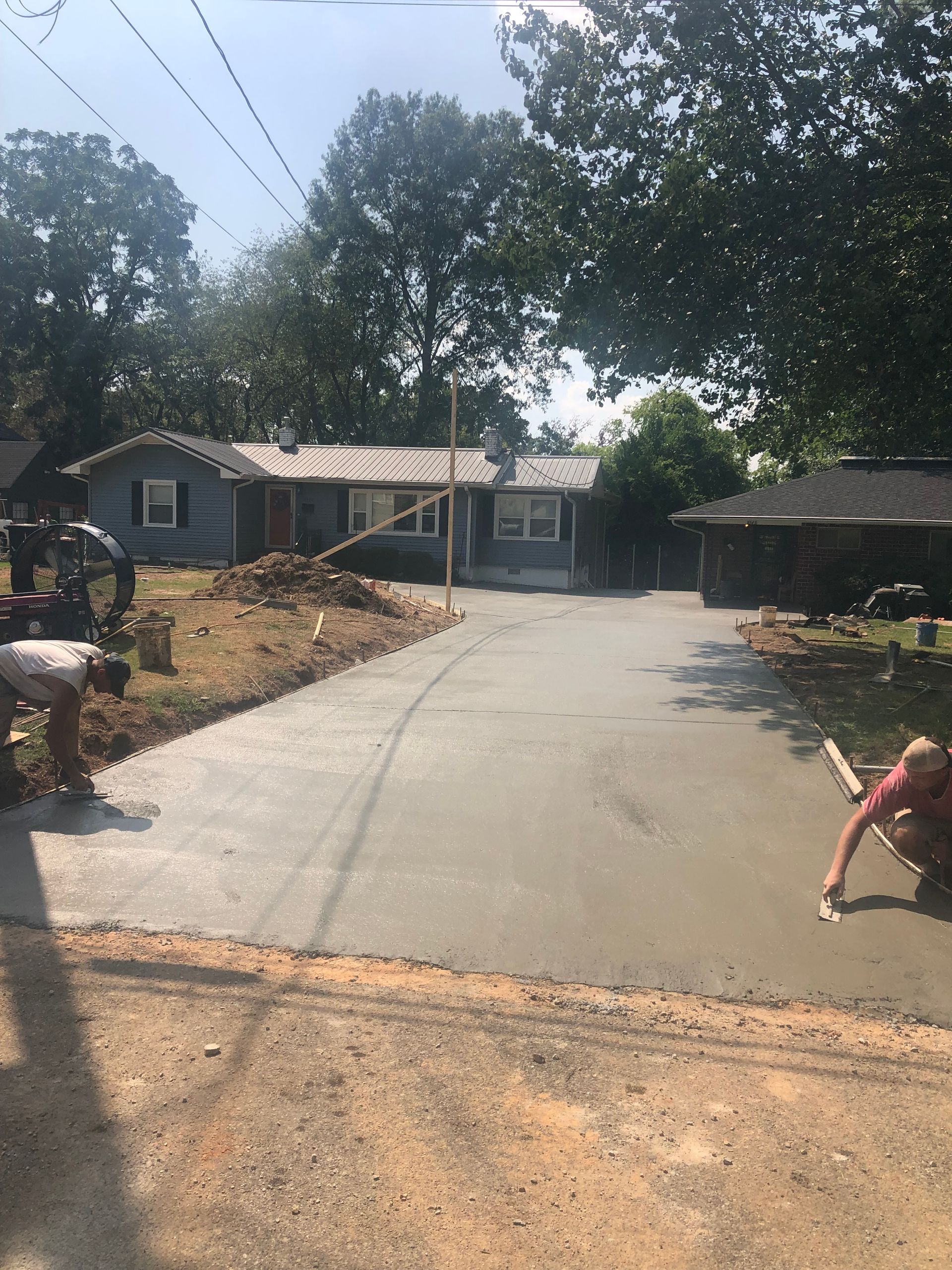 Workers smoothing wet concrete driveway in front of a blue house, trees in background.