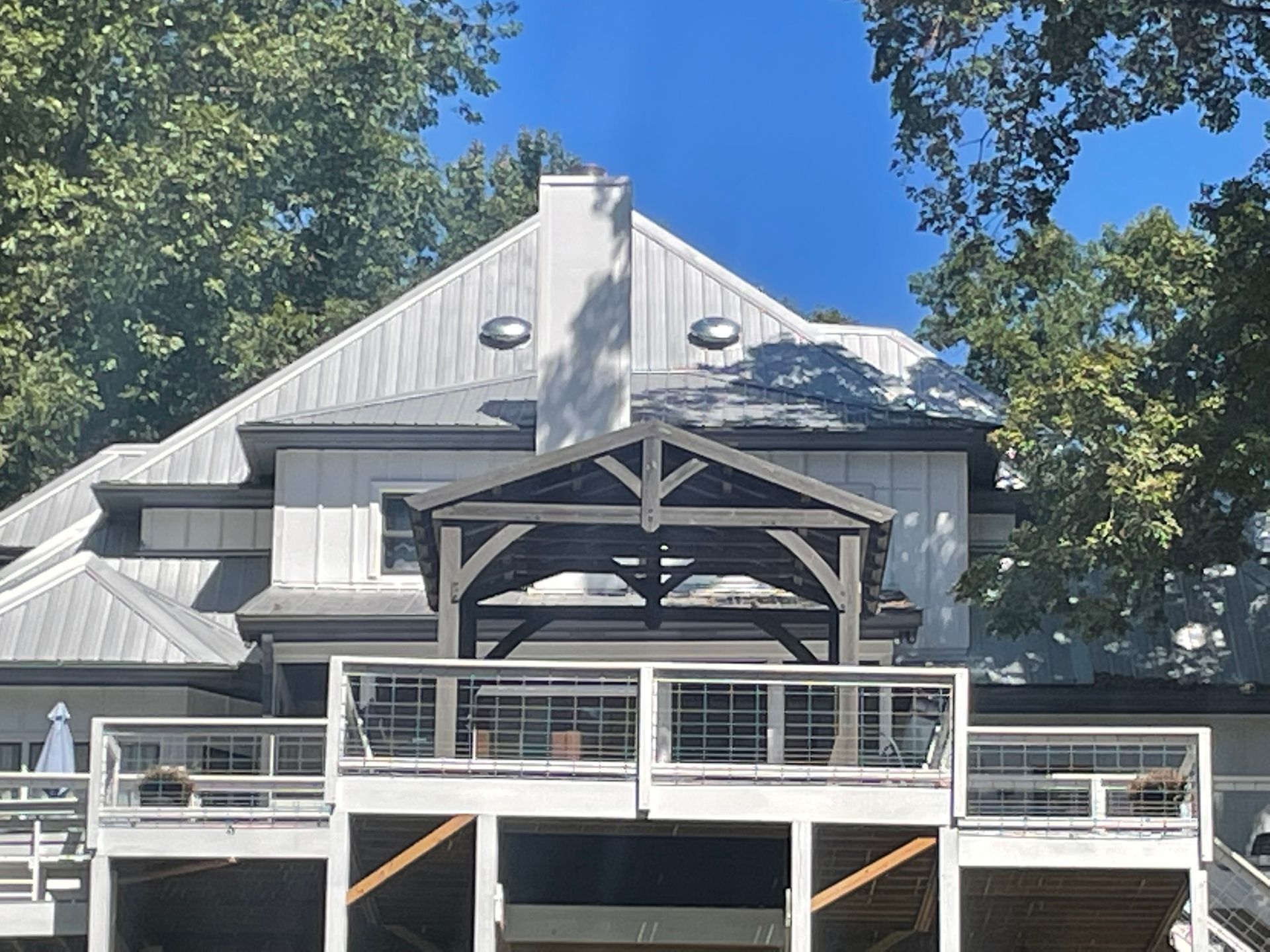 Back of a multi-level white house with a metal roof, deck, and pergola, surrounded by trees.