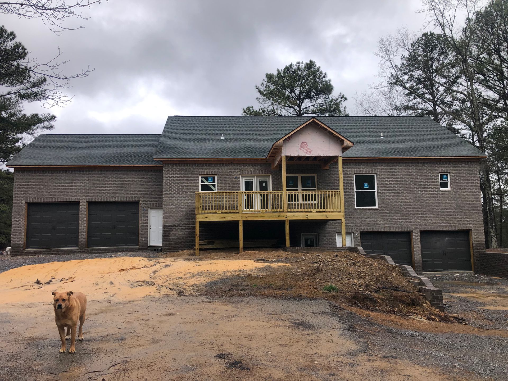 Brick house under construction with a deck, two garages, and a dog in the dirt yard.