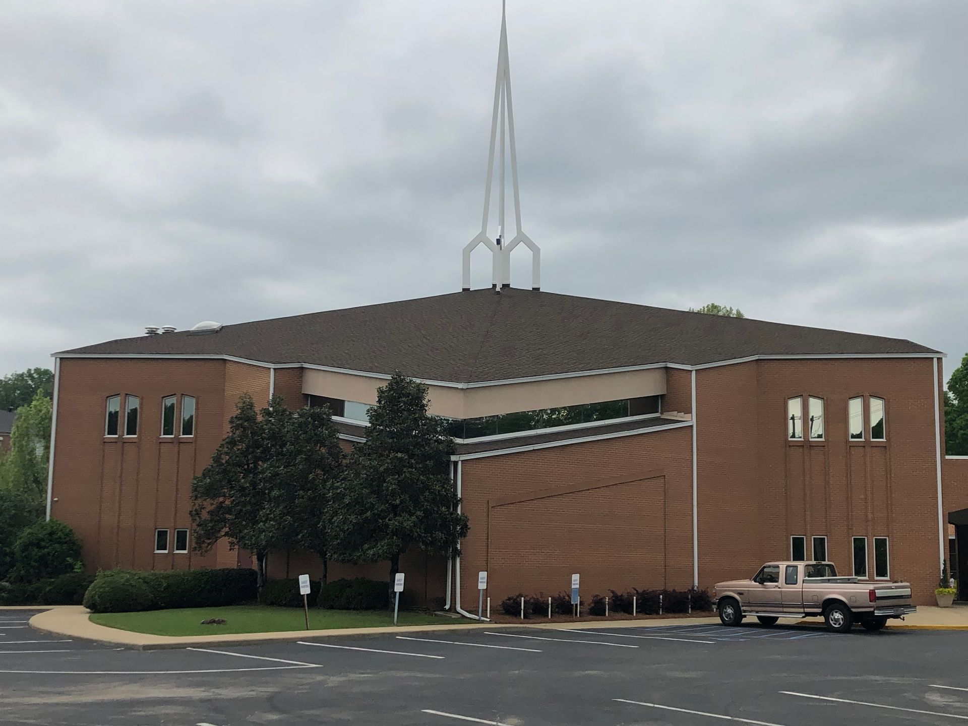 Brick church building with tall steeple, brown roof, and parked truck on a cloudy day.