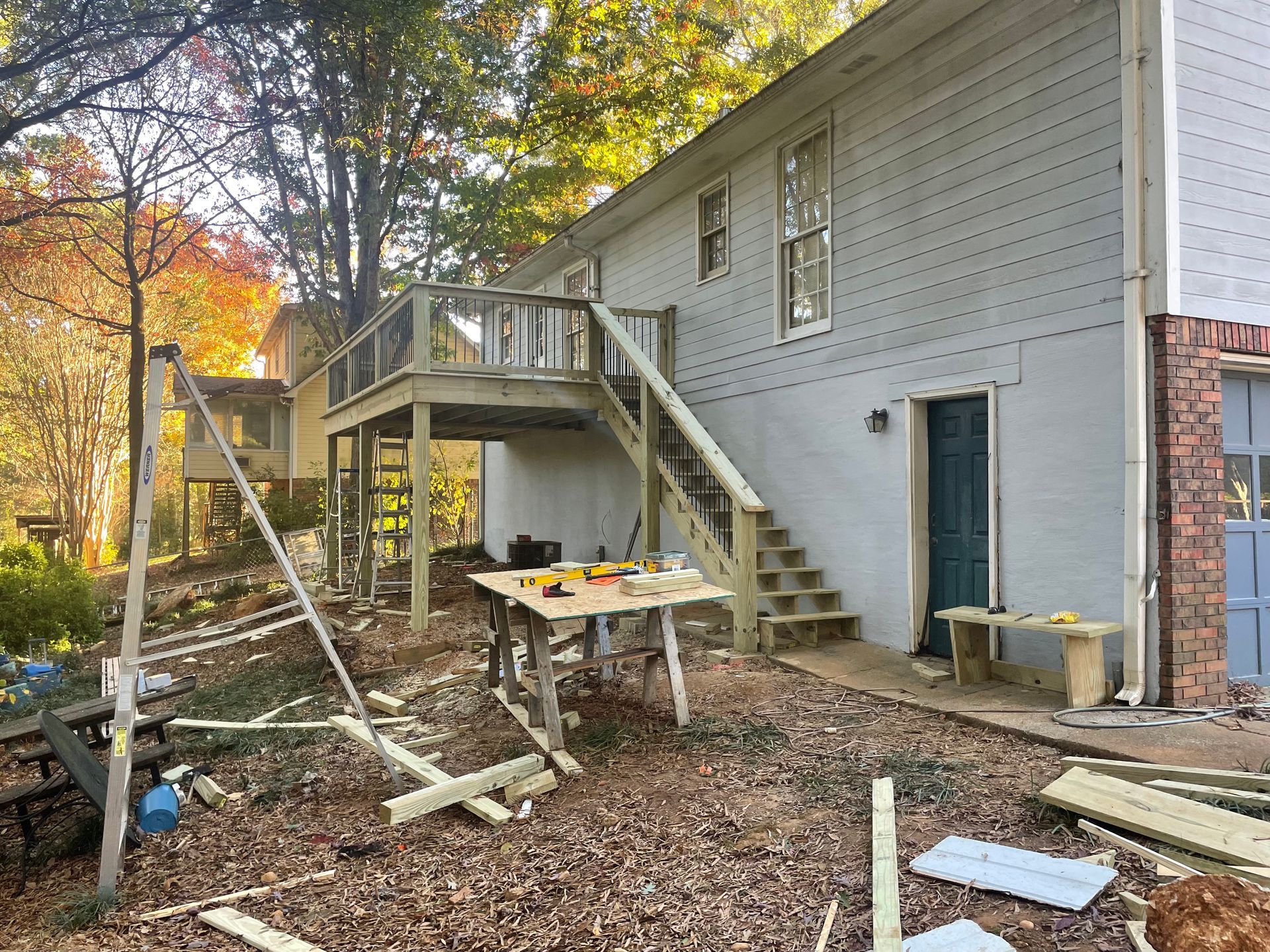 Exterior view of a two-story house with a wooden deck under construction; tools on a workbench, surrounded by fallen leaves.