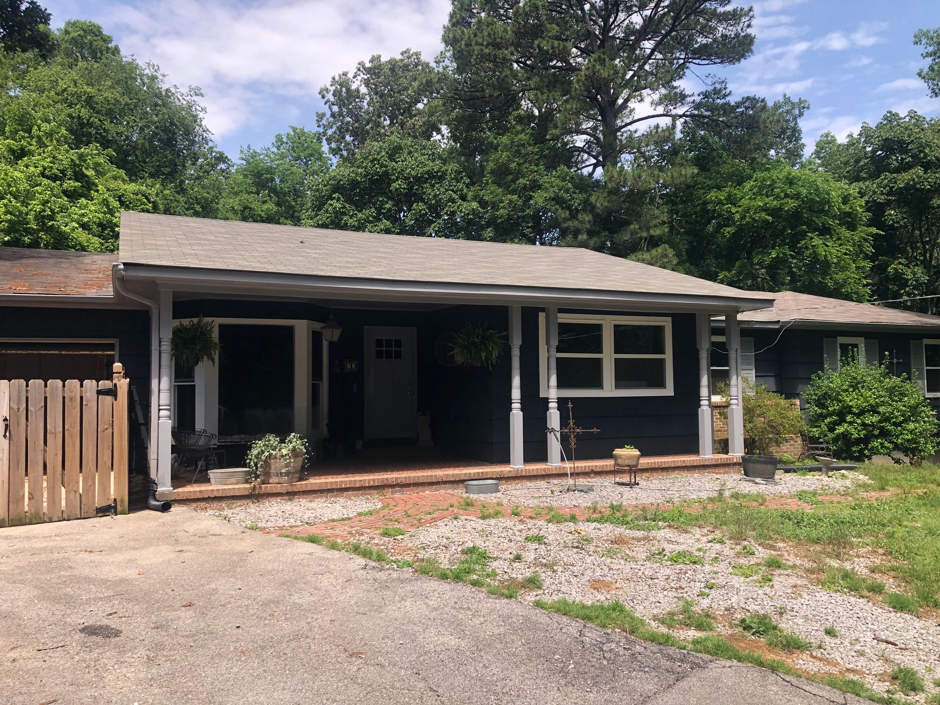 Dark blue house with a porch and brick accents, surrounded by greenery and a gravel driveway.