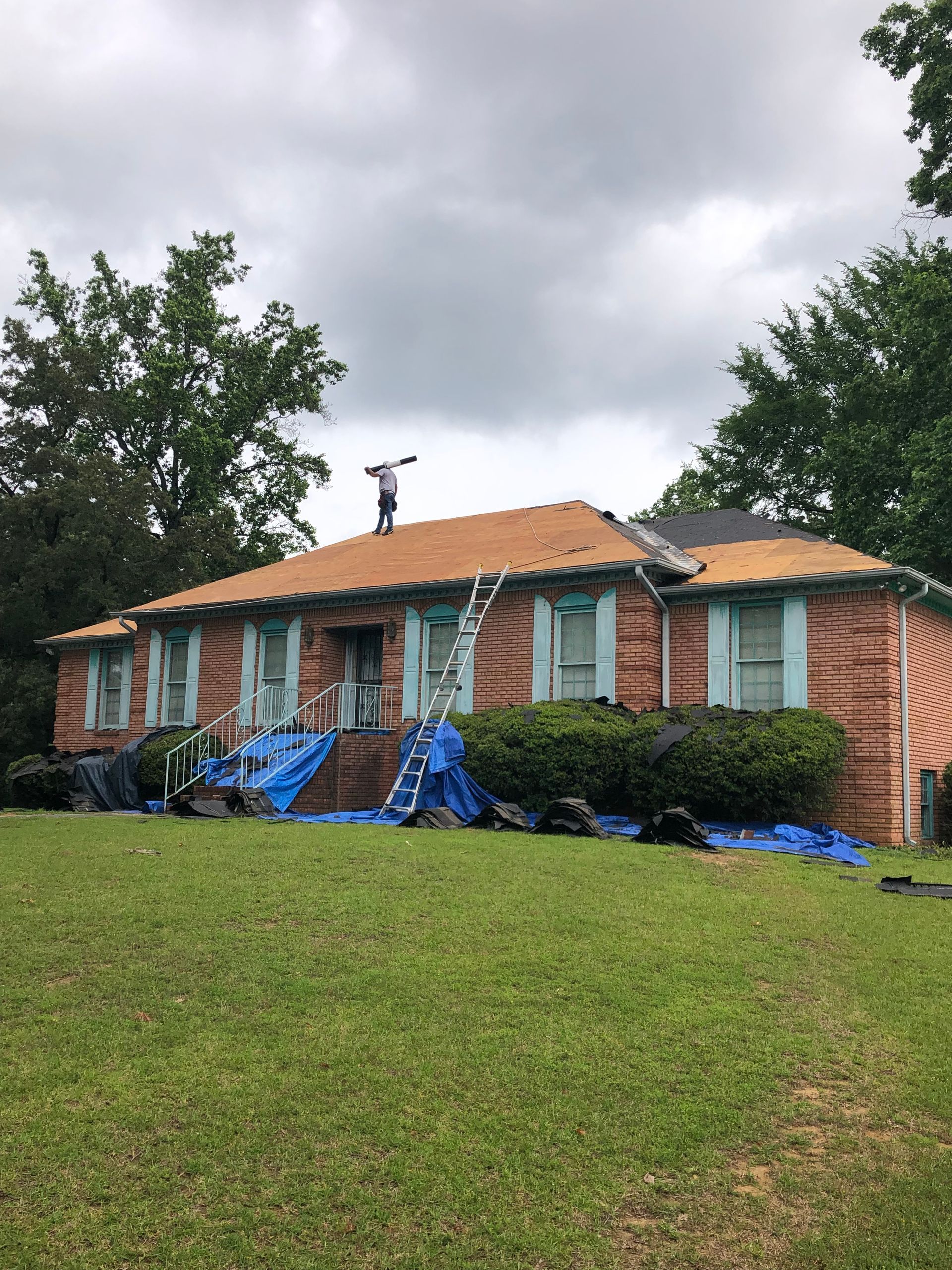 House with brick exterior, roof partially under construction. A worker on the roof, ladder present. Blue tarps cover the yard.