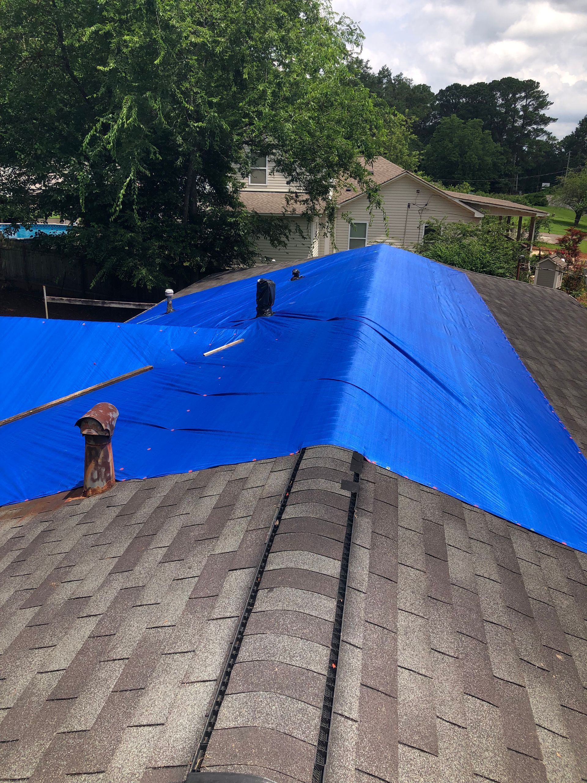 Blue tarp covering a roof with damaged shingles. A chimney is visible on the roof. Trees and a house in the background.