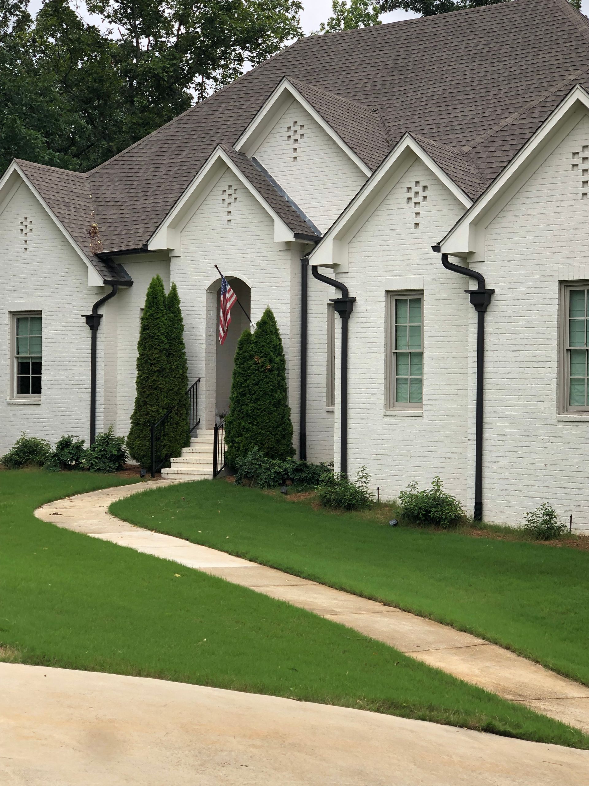 White brick house with dark roof and gutters. A winding driveway leads to the front door with an American flag.