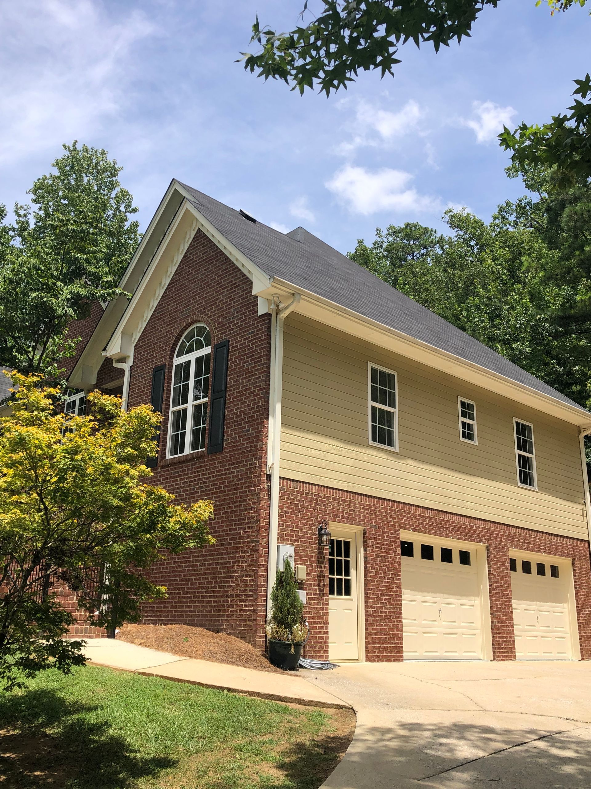 Two-story brick and tan house with a three-car garage, surrounded by trees under a blue sky.