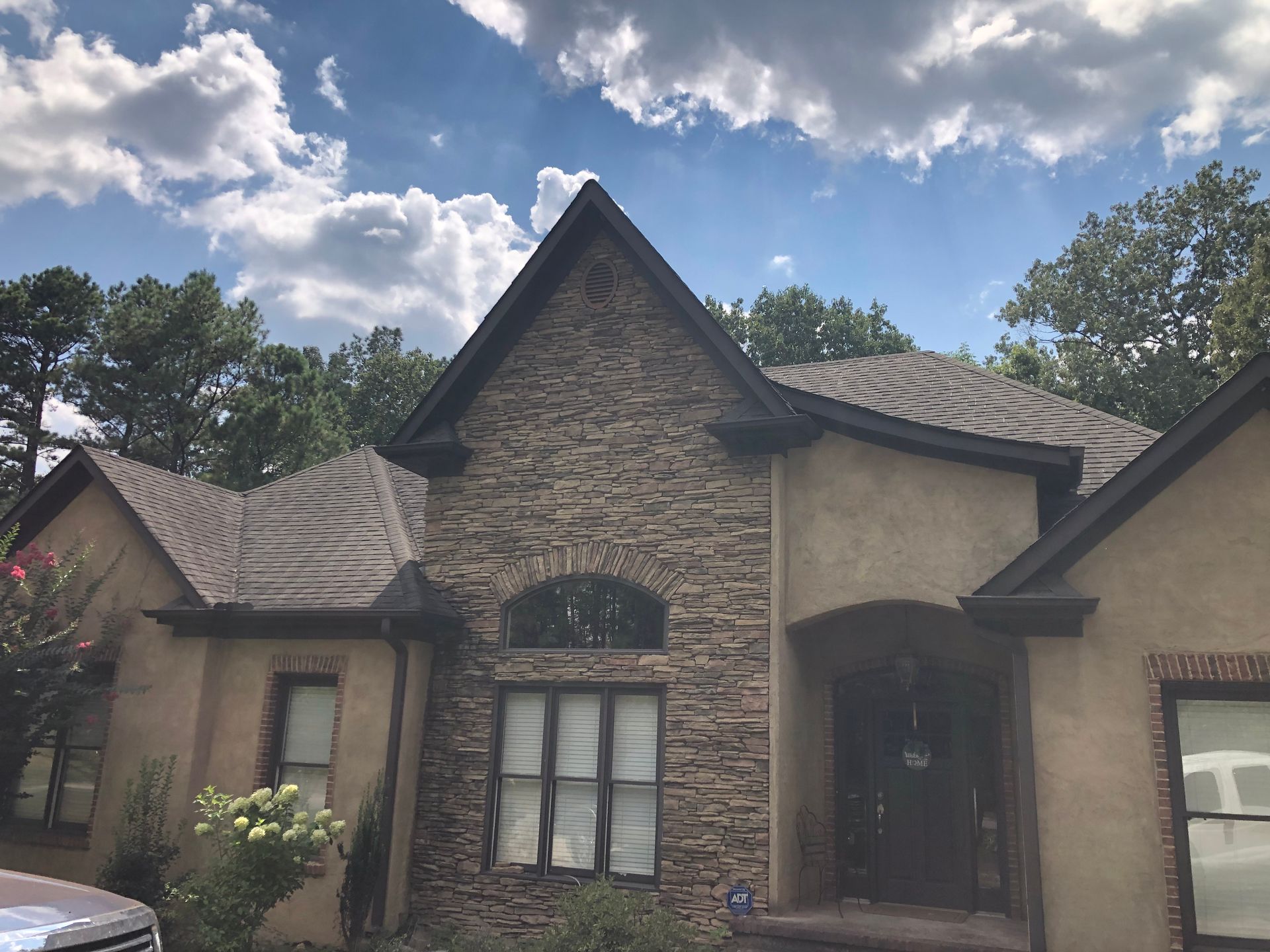 Stone-faced house with brown roof and trim. Trees and cloudy sky in the background.
