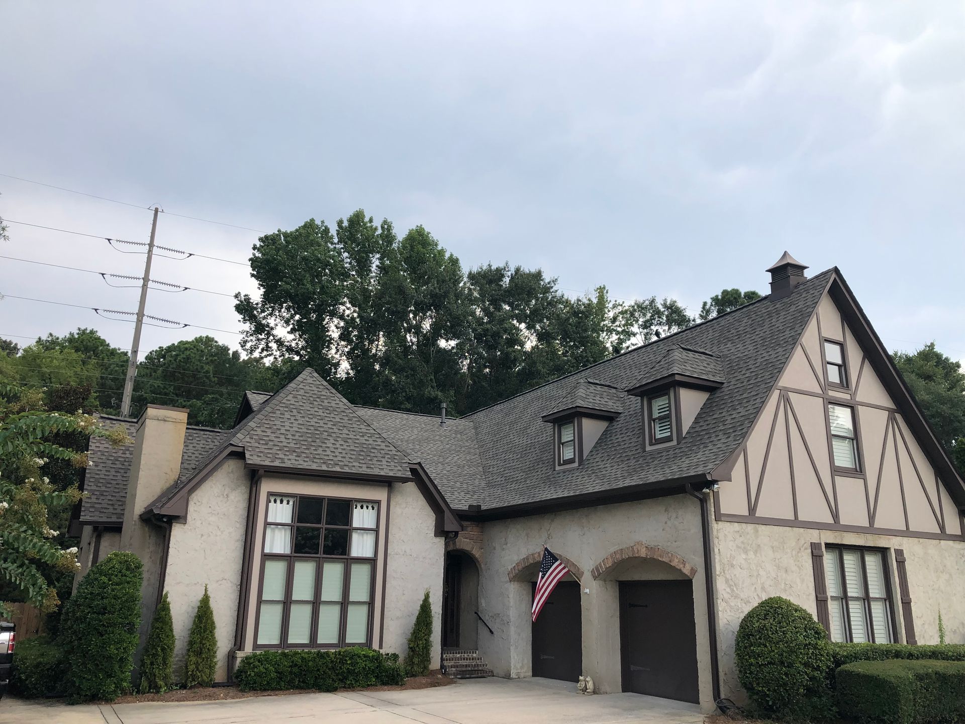Tudor-style house with tan stucco, dark roof, decorative beams, and garage. An American flag hangs on the side.