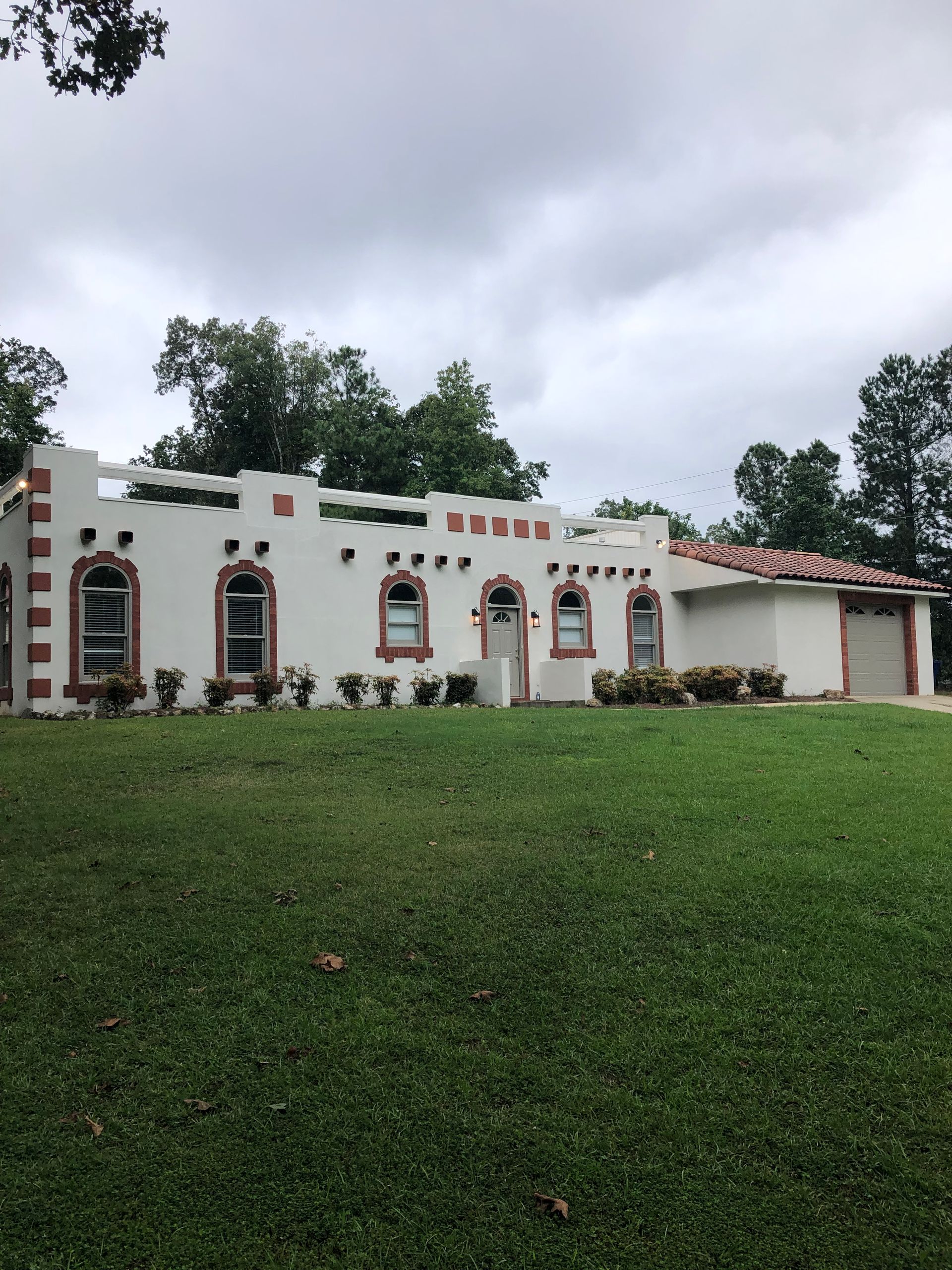 White stucco home with red trim, arched windows, and a garage, set on a green lawn under a cloudy sky.