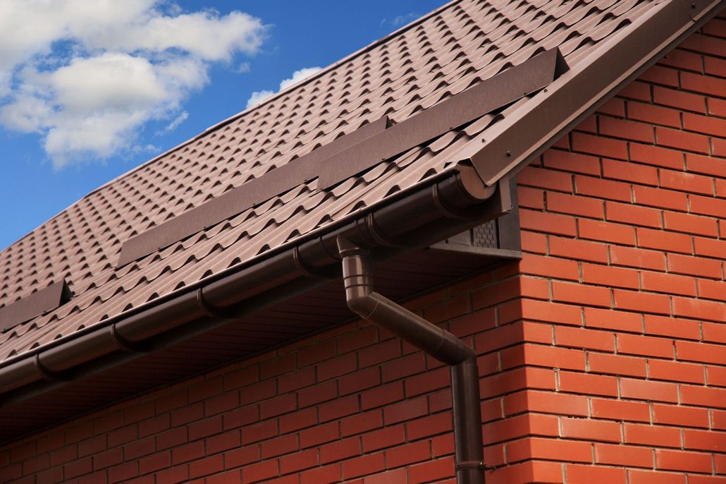 Brown metal roof with matching gutters and a downspout attached to a red brick house under a blue sky with white clouds.
