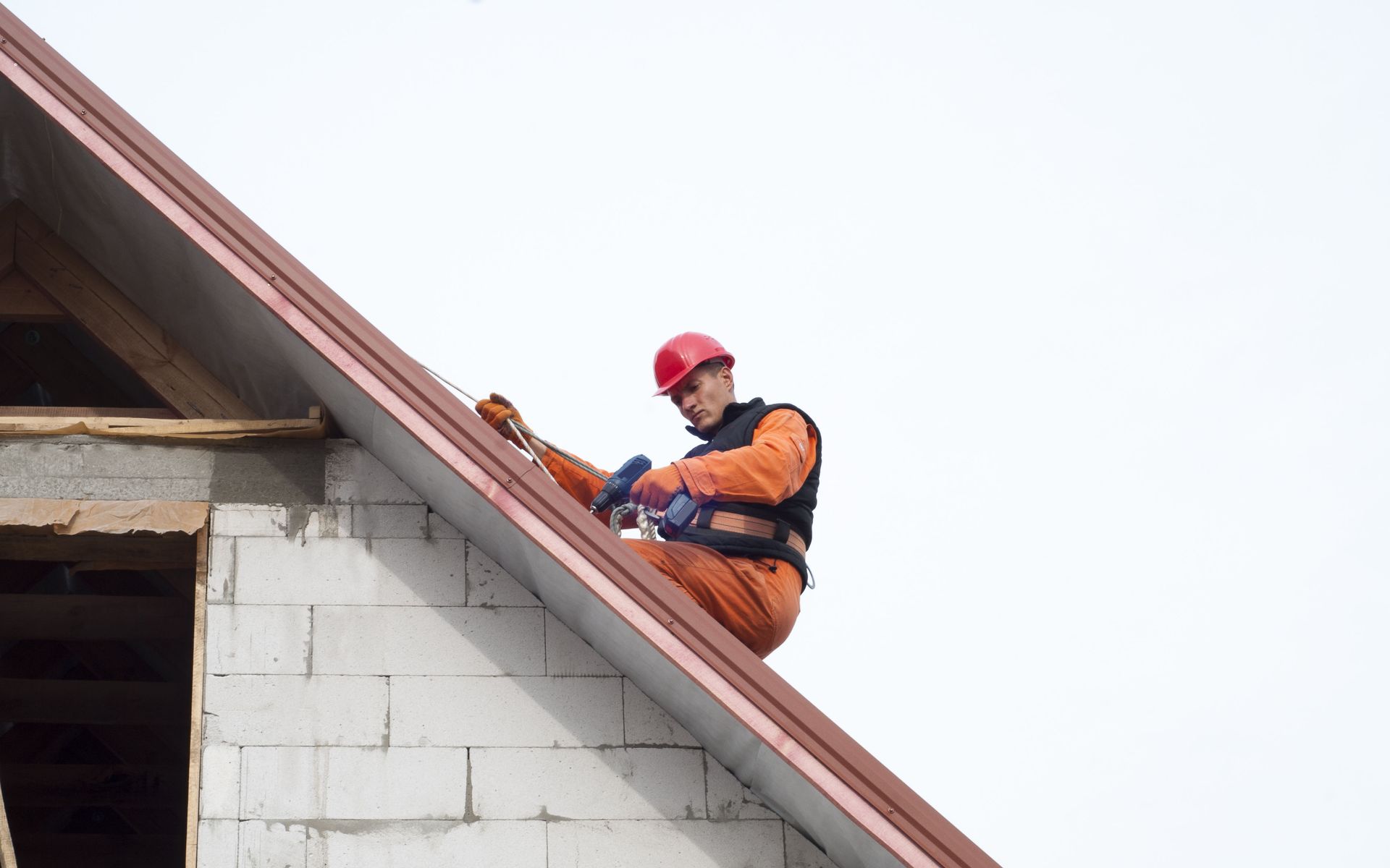 A construction worker in an orange uniform and red hard hat works on the edge of a sloped roof.