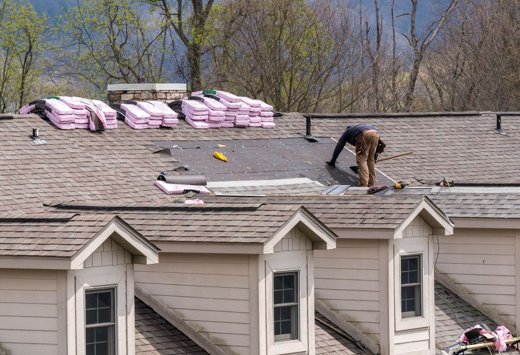A worker in brown pants repairs a shingled roof with pink insulation boards stacked nearby on a sunny day.