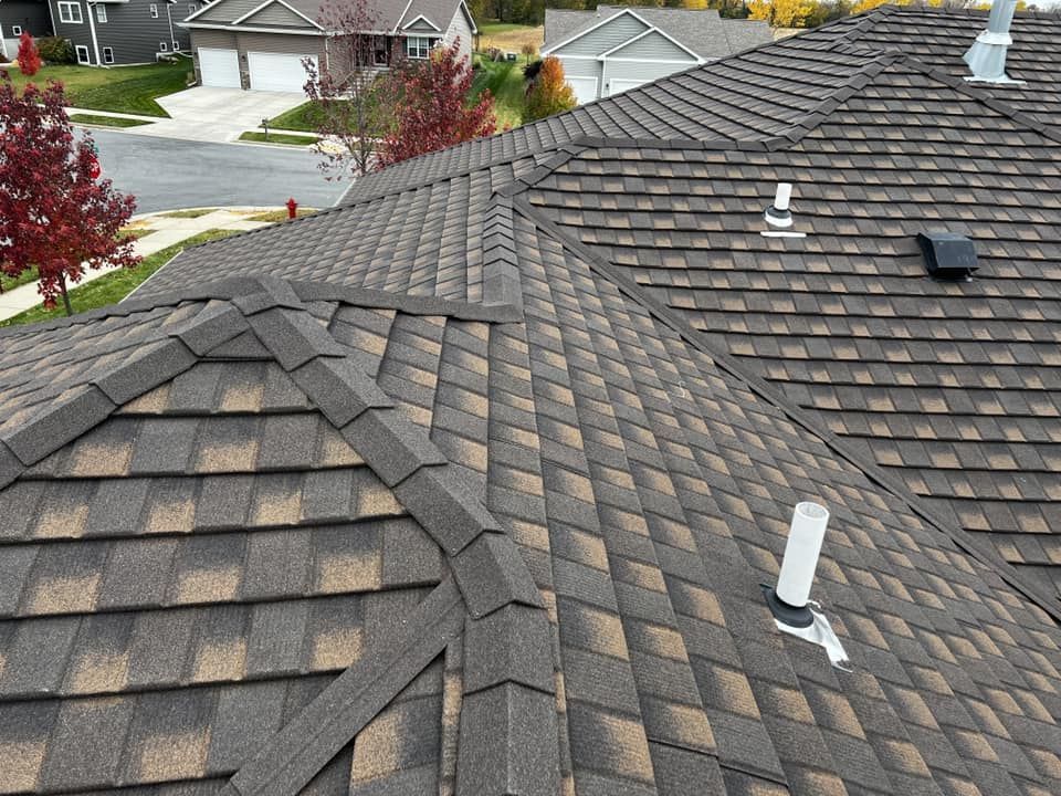 A close-up of a brown and tan shingle roof with several vents and a chimney in a suburban setting.