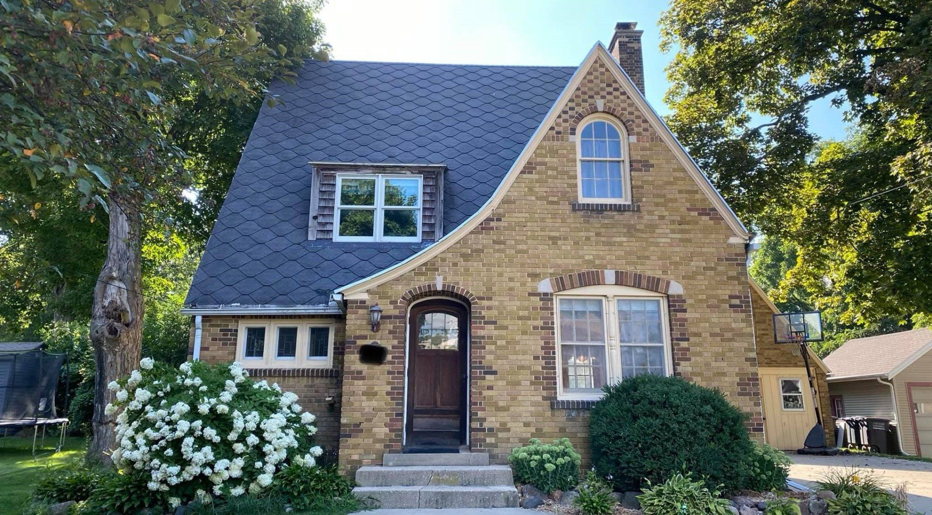 Tan brick house with a dark roof, arched doorway, and small windows. Green bushes and trees frame the house.