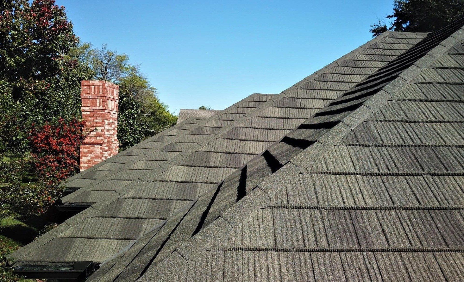 Close-up of a textured dark gray roof with a red brick chimney and trees under a blue sky.