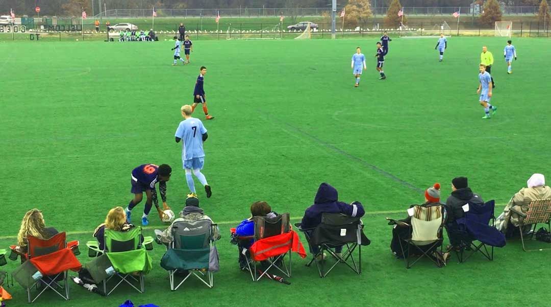 Soccer game on green field; players in light blue and dark uniforms. Spectators in chairs watch.