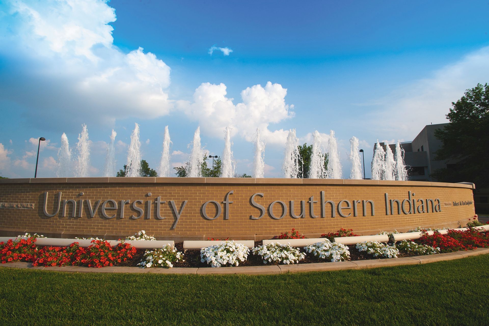 University of Southern Indiana sign with fountain and flowers in front of a building under a cloudy sky.