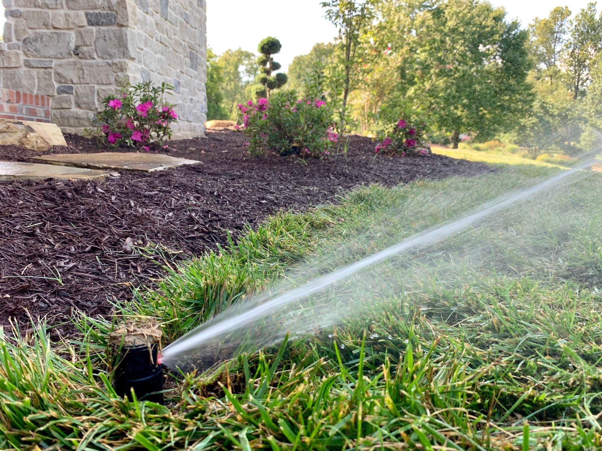 Sprinkler watering green grass in a landscaped yard with flowers and a stone building in the background.