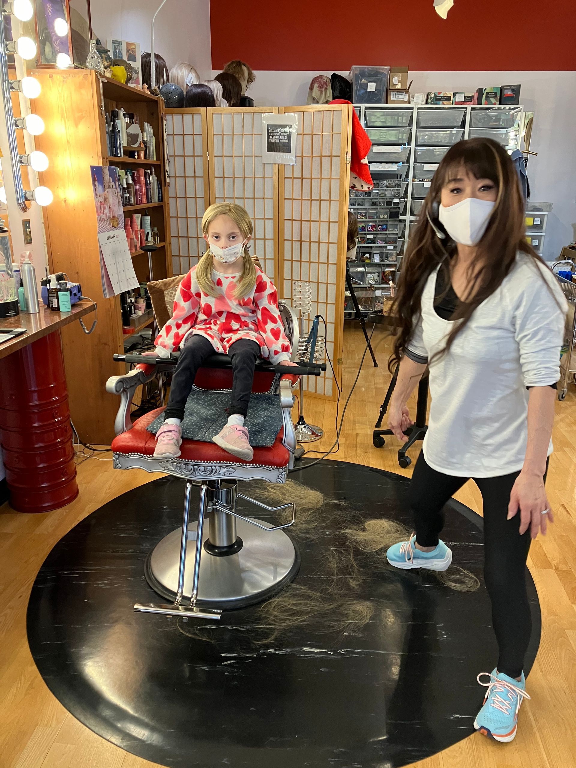 A woman wearing a mask is standing next to a little girl in a barber chair.
