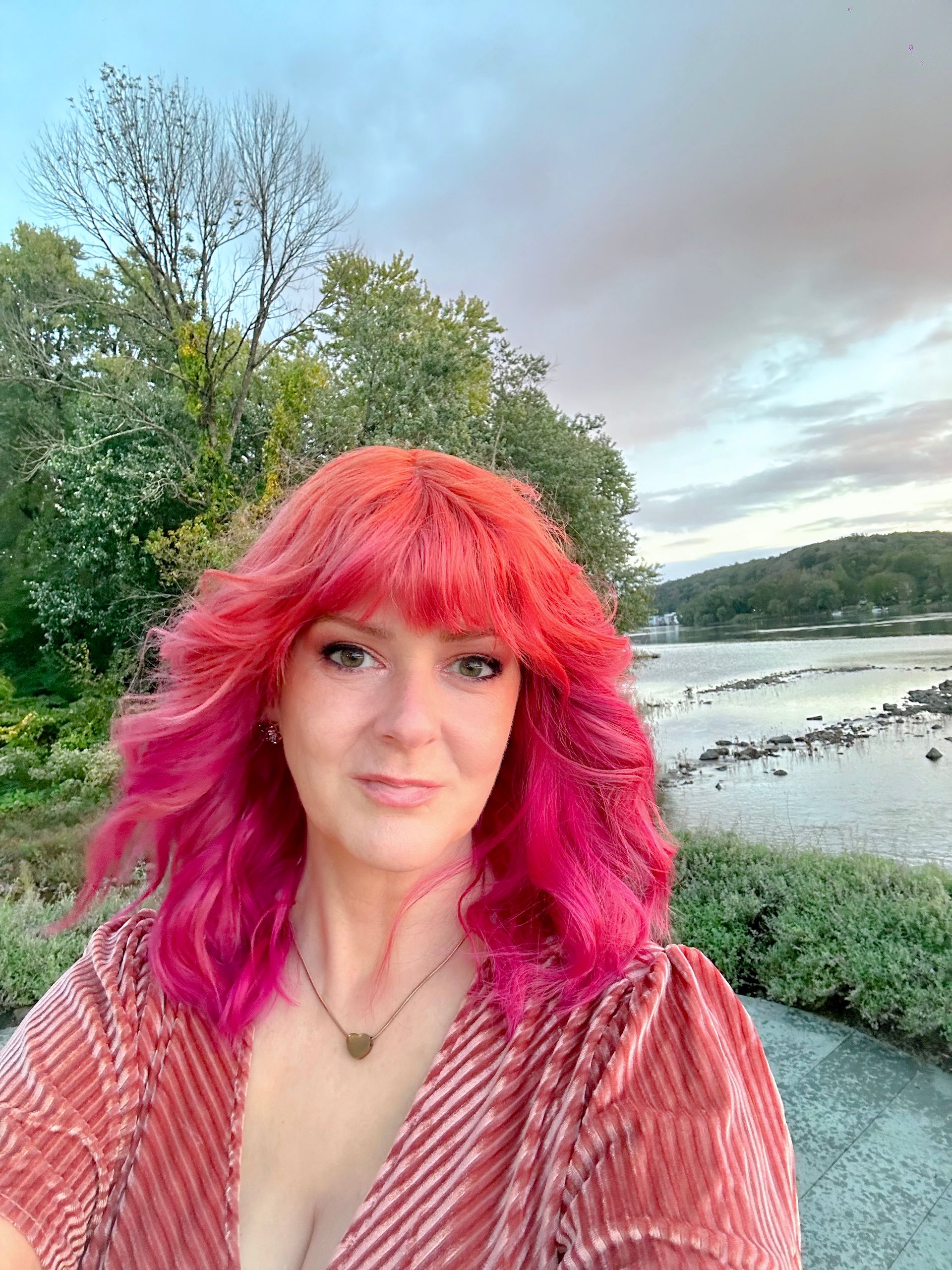 A woman with pink hair is standing in front of a body of water.
