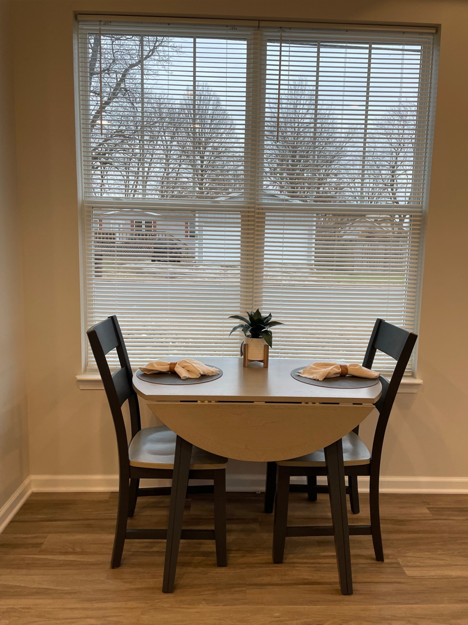 Dining table with two chairs in front of a window with blinds. Plates on table; plant in the center.